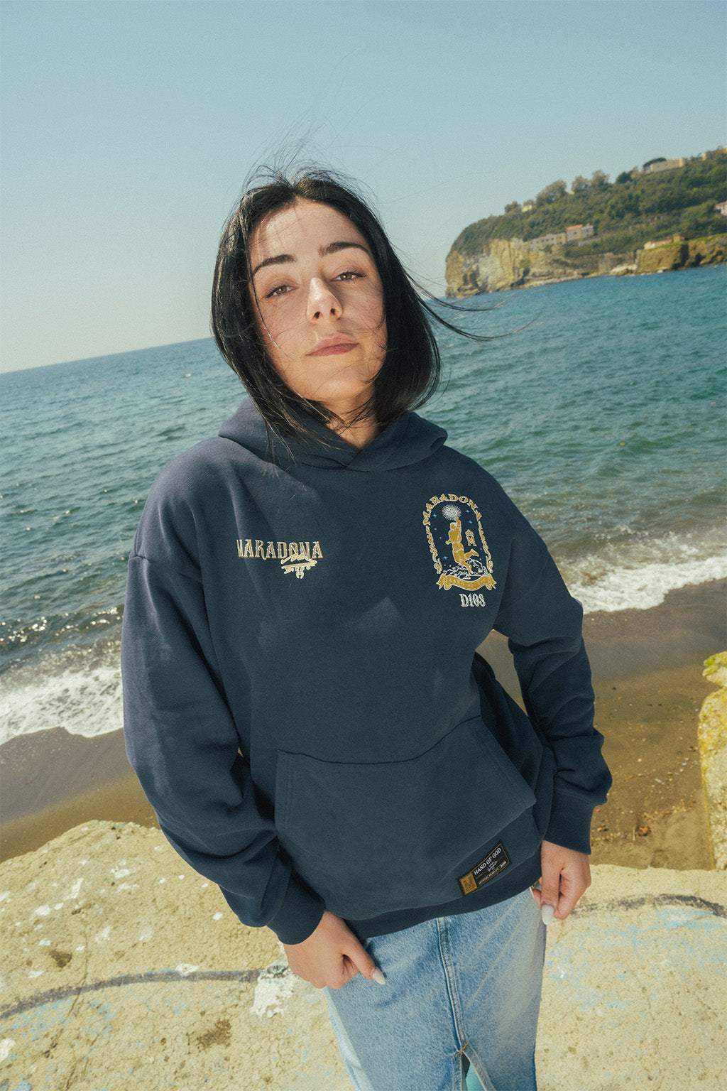 Woman wearing a navy Maradona hoodie while standing on a stone shore with ocean in background