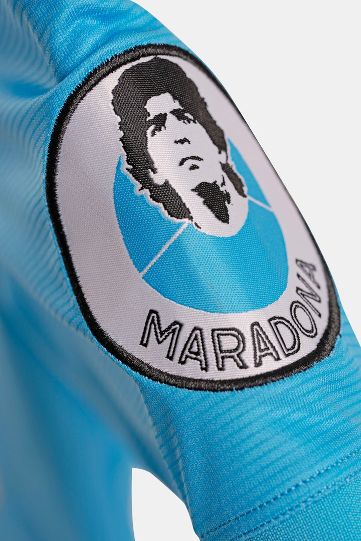 Close-up of a light blue soccer jersey sleeve featuring a circular embroidered patch with a black-and-white portrait and the name 'MARADONA' stitched along the bottom.