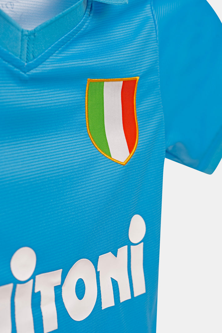Close-up of a light blue soccer jersey with a shield crest featuring green, white, and red vertical stripes outlined in gold, and a large white sponsor logo across the front.