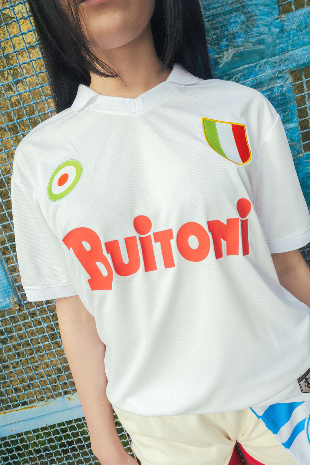 Close up woman wearing a white Napoli replica jersey against a textured blue grated wall.
