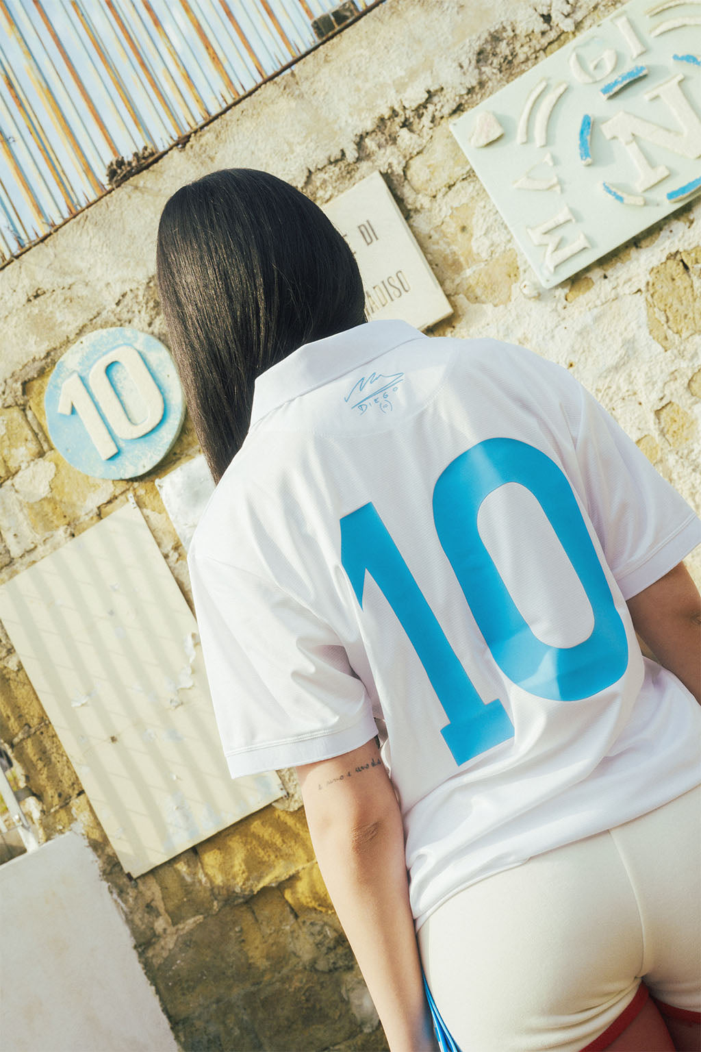 Woman wearing a white Napoli replica jersey with the number 10 on the back, standing against a textured wall.