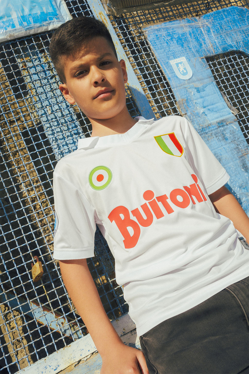 Young boy wearing a white Napoli replica jersey against a grated wall.