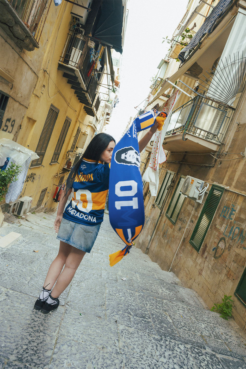 Woman holding a Boca inspired Maradona scarf in an urban alleyway with buildings on either side.