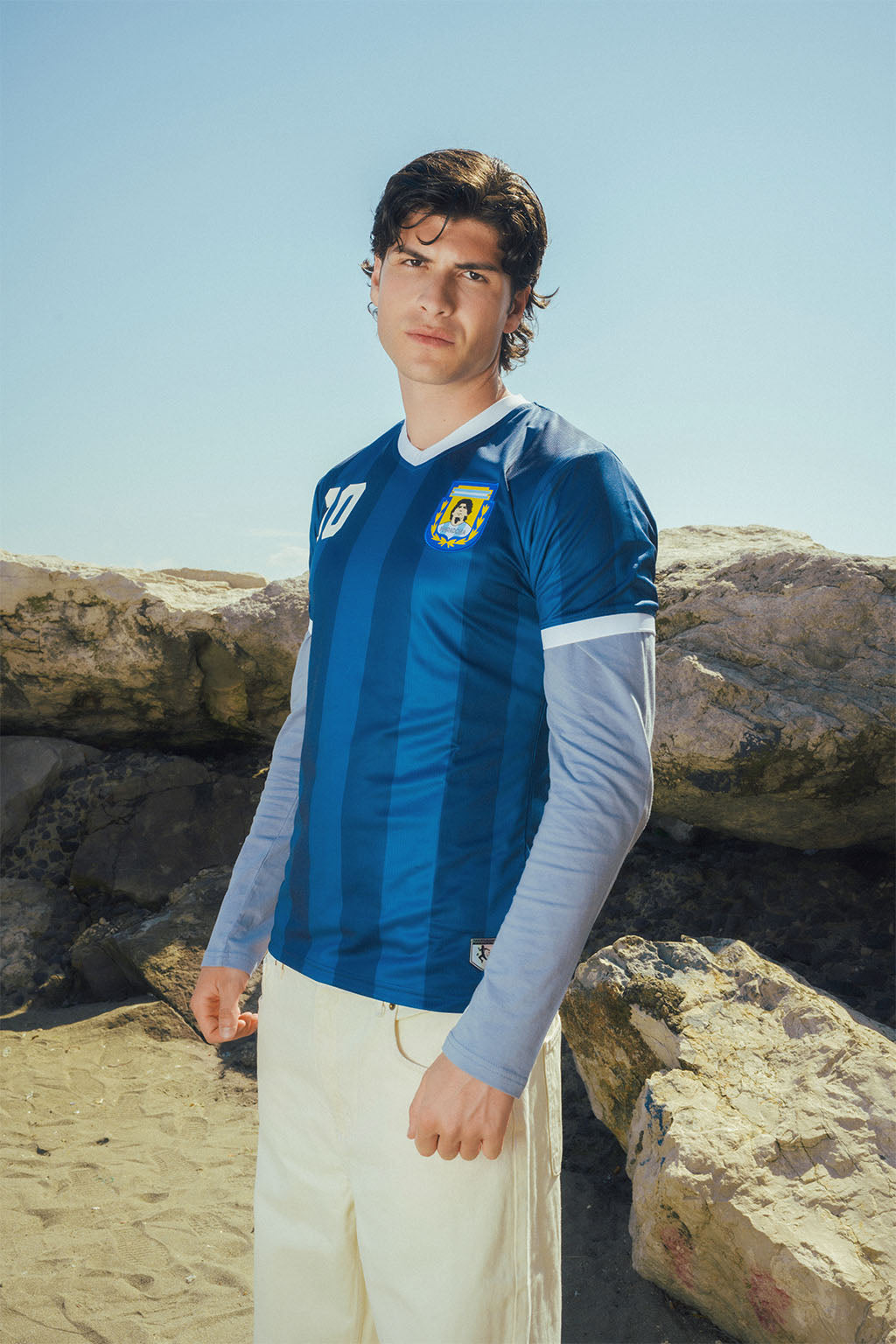 Man wearing a blue Argentina Away sports jersey with a Maradona logo, standing on a beach.