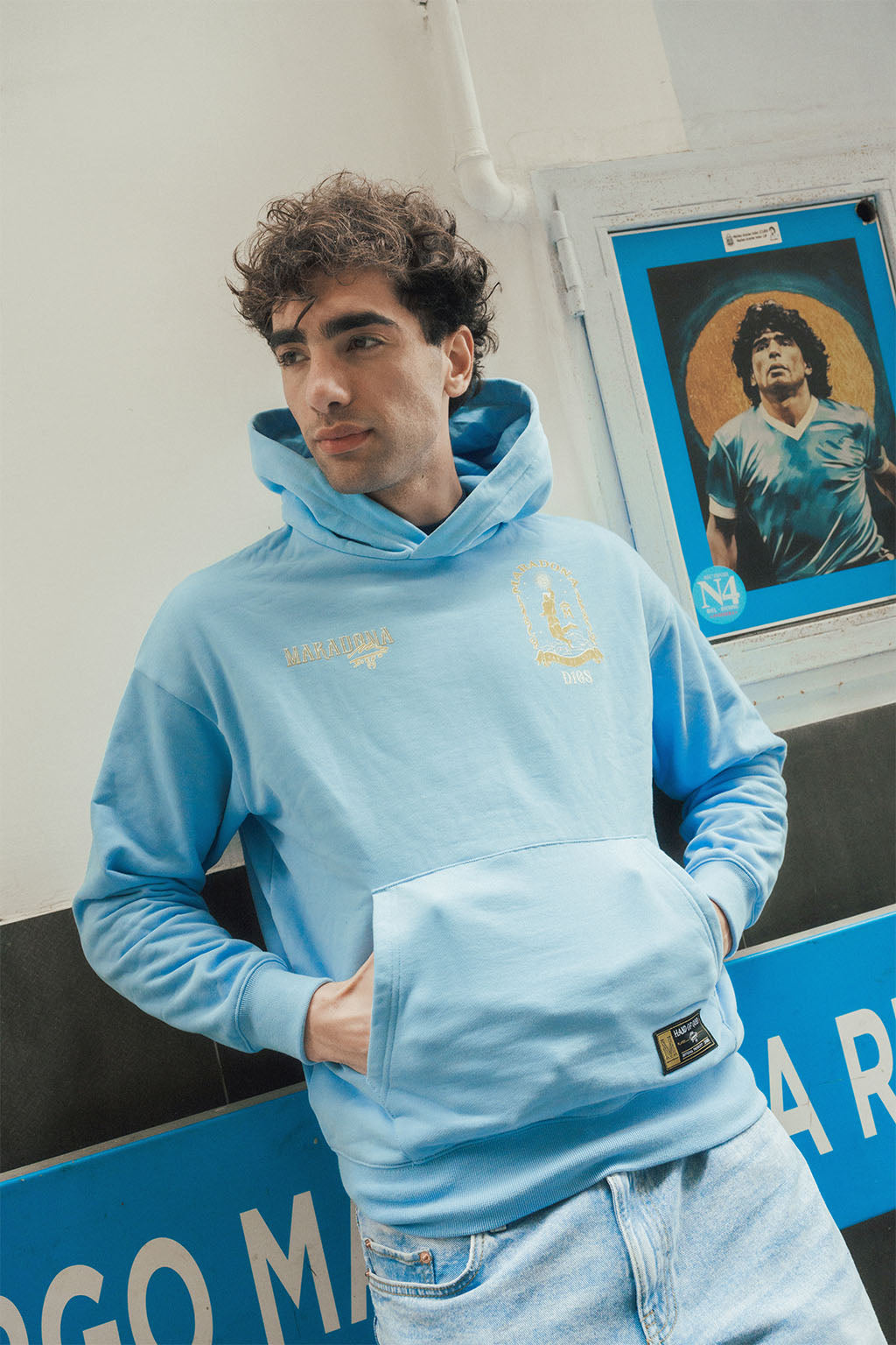 Man wearing a light blue hoodie with a gold Maradona logo, standing in front of a wall with a Diego poster.