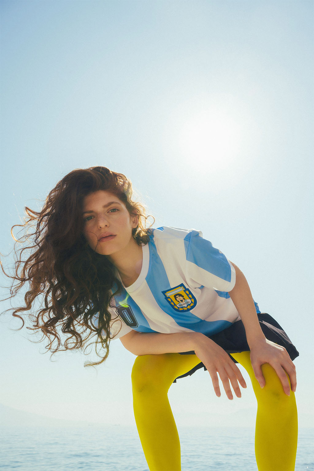 Woman wearing a blue and white striped Argentina home jersey with a Maradona logo, yellow stockings, standing on beach against a clear blue sky.