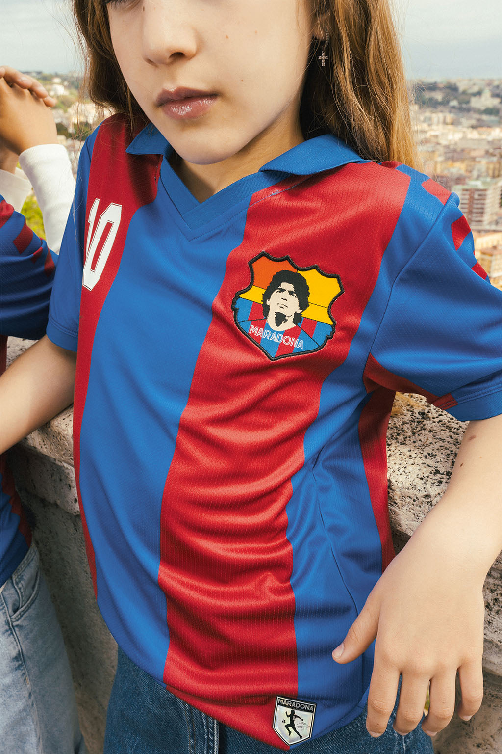 Child wearing a red and blue Barcelona replica jersey with Maradona crest, standing outdoors.