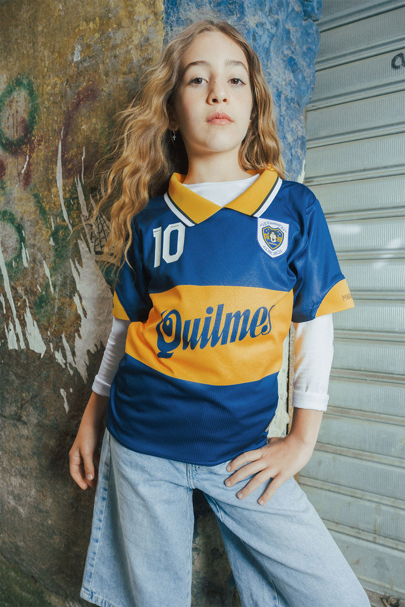 Child wearing a blue and yellow Boca replica jersey with 'Quilmes' against a textured urban wall.