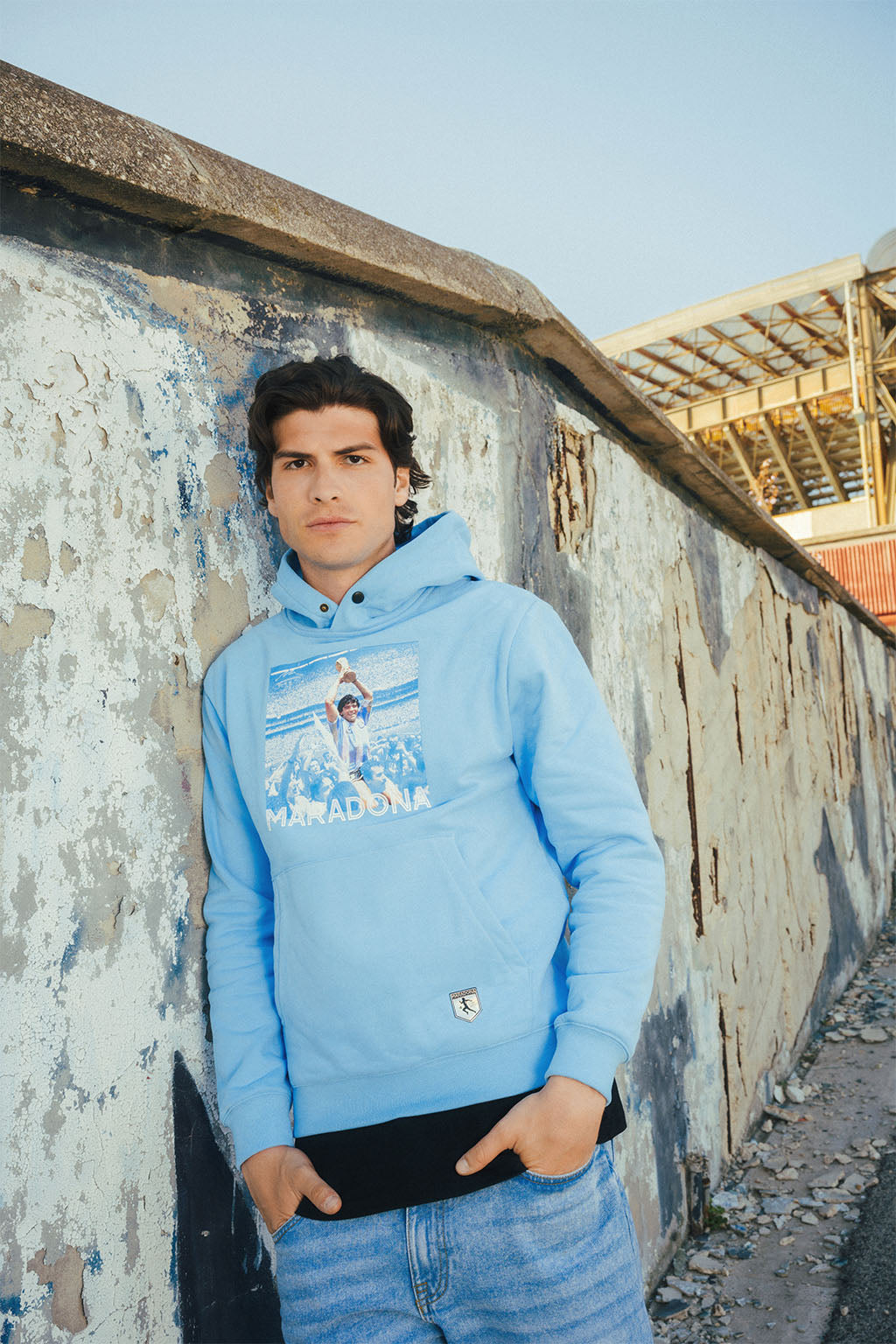 Main wearing a blue hoodie with Maradona graphic print leaning against a wall with graffiti.