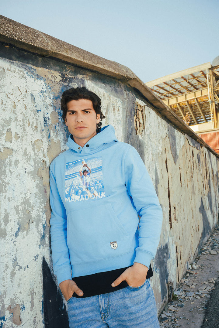 Main wearing a blue hoodie with Maradona graphic print leaning against a wall with graffiti.