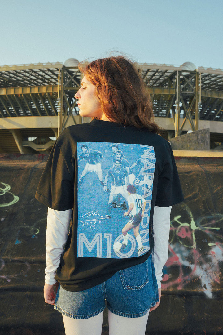 Woman wearing a black t-shirt with a graphic Maradona 86 design, standing in outside stadium with graffiti on the wall