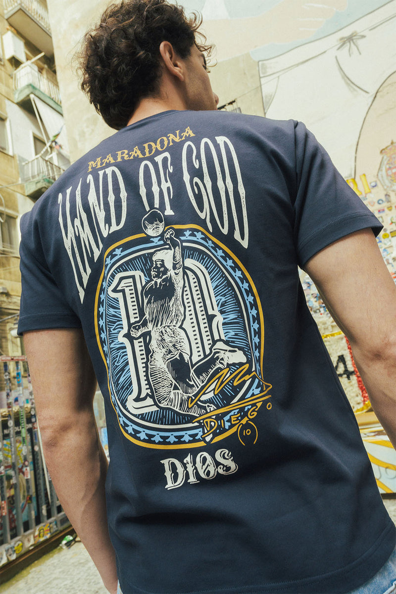 Man wearing a navy blue tee with graphic Hand of God design on the back, standing in an urban setting.