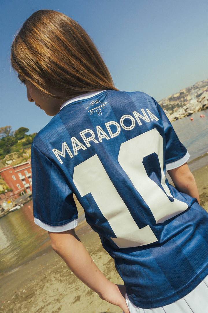 Young girl wearing a blue sports jersey with 'Maradona' and number '10' on back at a beach.