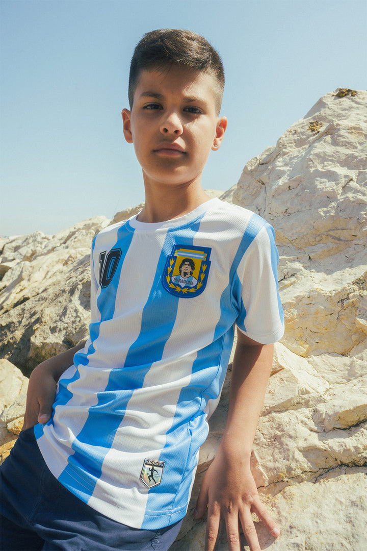 Child wearing a blue and white striped jersey with a Maradona crest, standing on beach rocks.