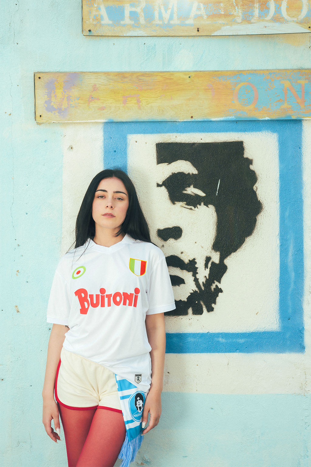 Woman wearing a replica Napoli jersey in front of a wall with a Maradona portrait and faded signs.