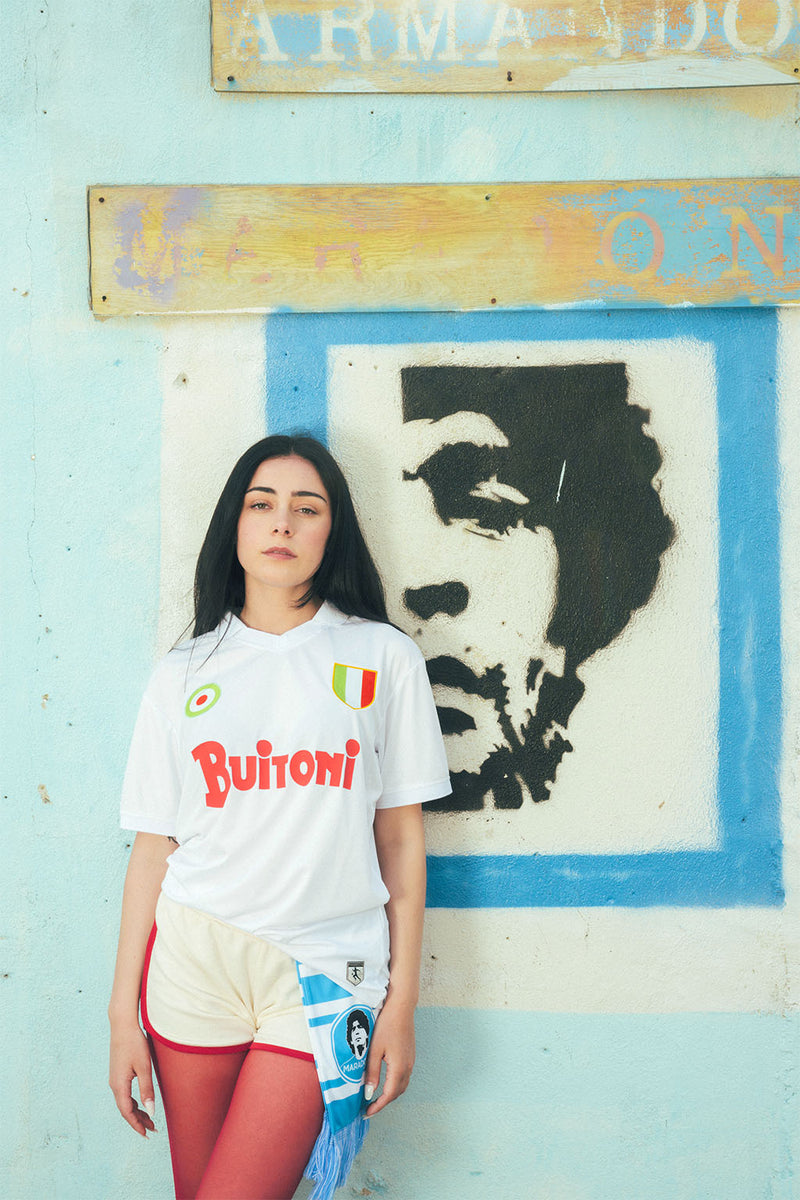 Woman wearing a replica Napoli jersey in front of a wall with a Maradona portrait and faded signs.