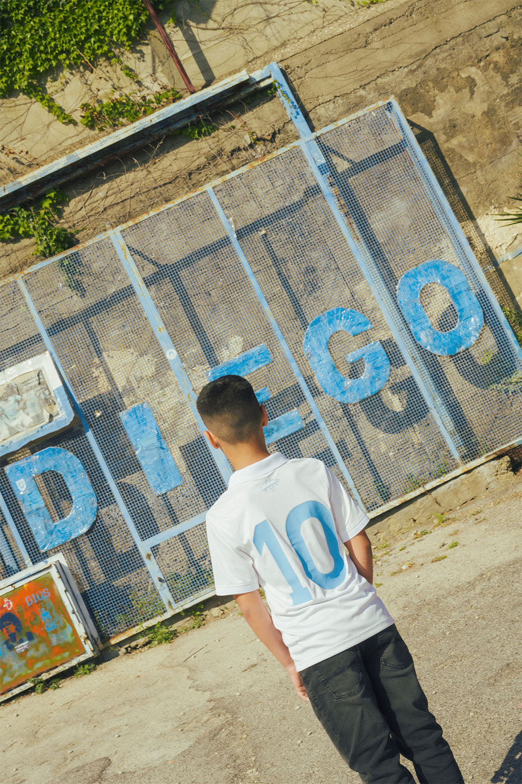 boy wearing a white Napoli replica jersey with blue numbers standing in front of a decorative metal gate with 'DIEGO' written on it.