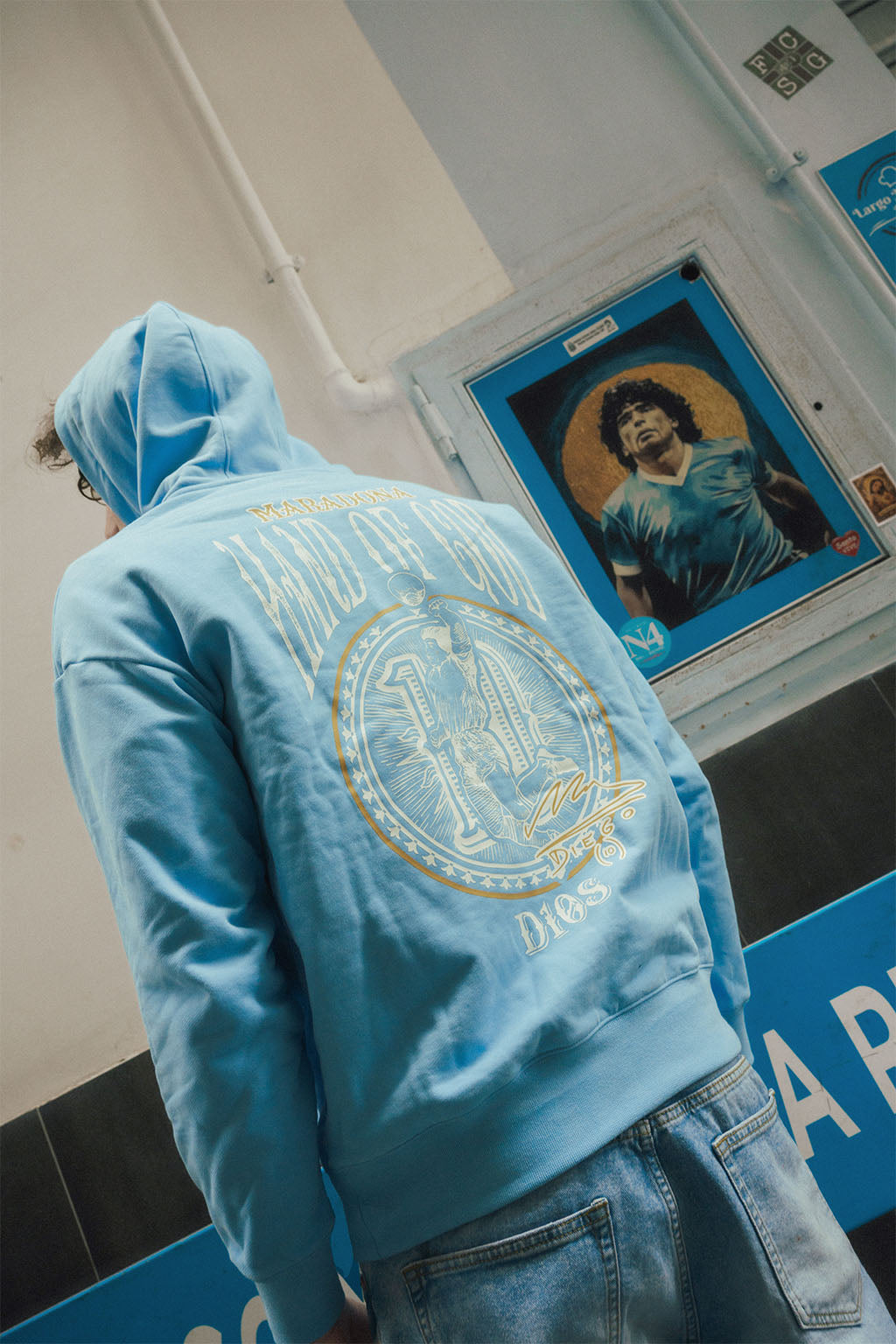 Man facing away wearing a blue oversize hoodie with a Hand of God graphic design, standing in front of a Diego Maradona poster