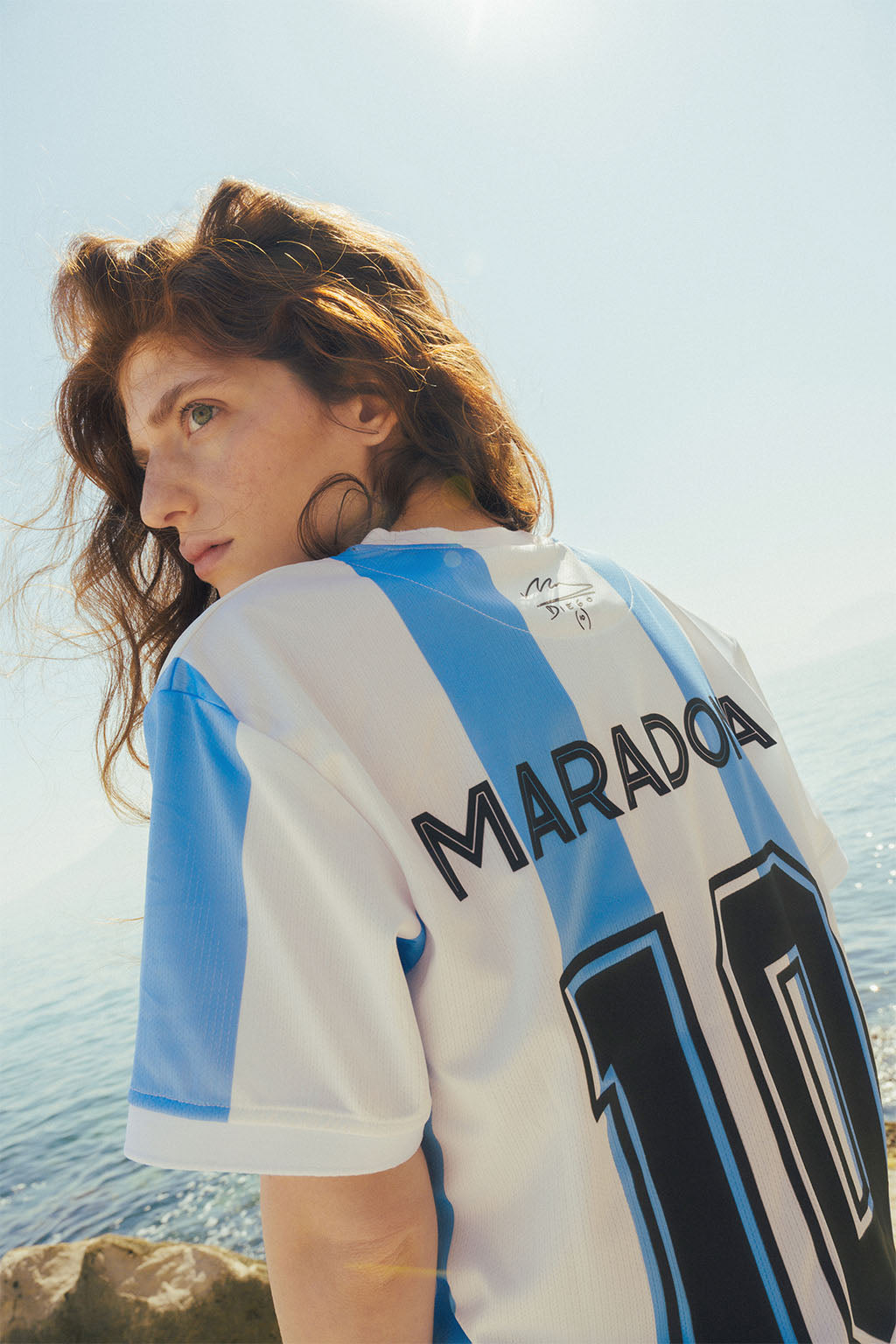 Woman wearing a Argentina sports jersey with 'Maradona' and number '10' standing on beach against a clear sky.
