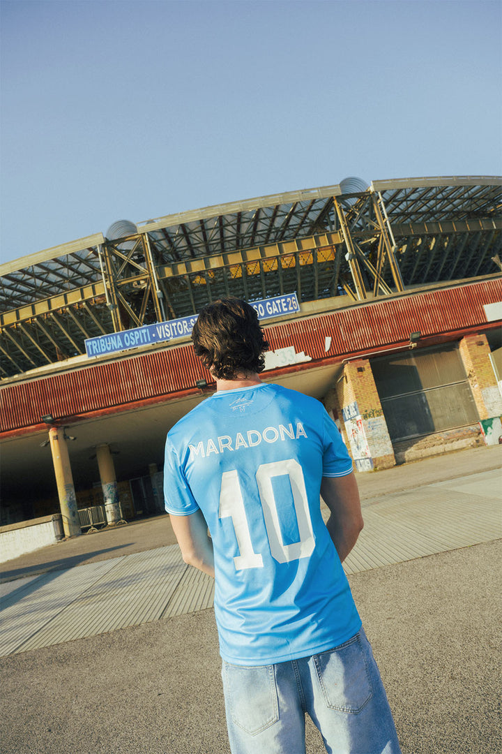Man wearing a replica Napoli blue sports jersey with 'Maradona' and number '10' in front of an old stadium.
