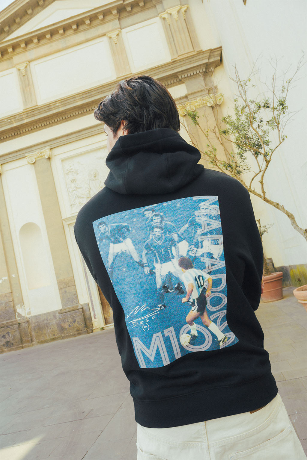Man wearing a black hoodie with a graphic Maradona design on the back, standing in front of an architectural building.