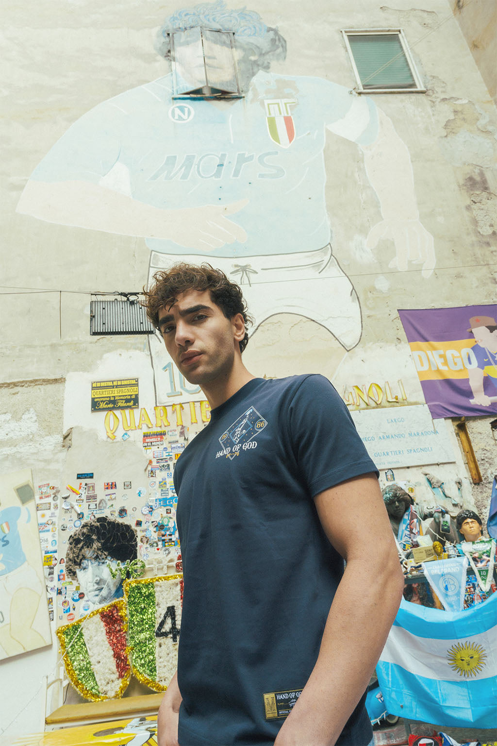 Man standing in front of a Maradona collage wall wearing navy tee with Hand of God designs