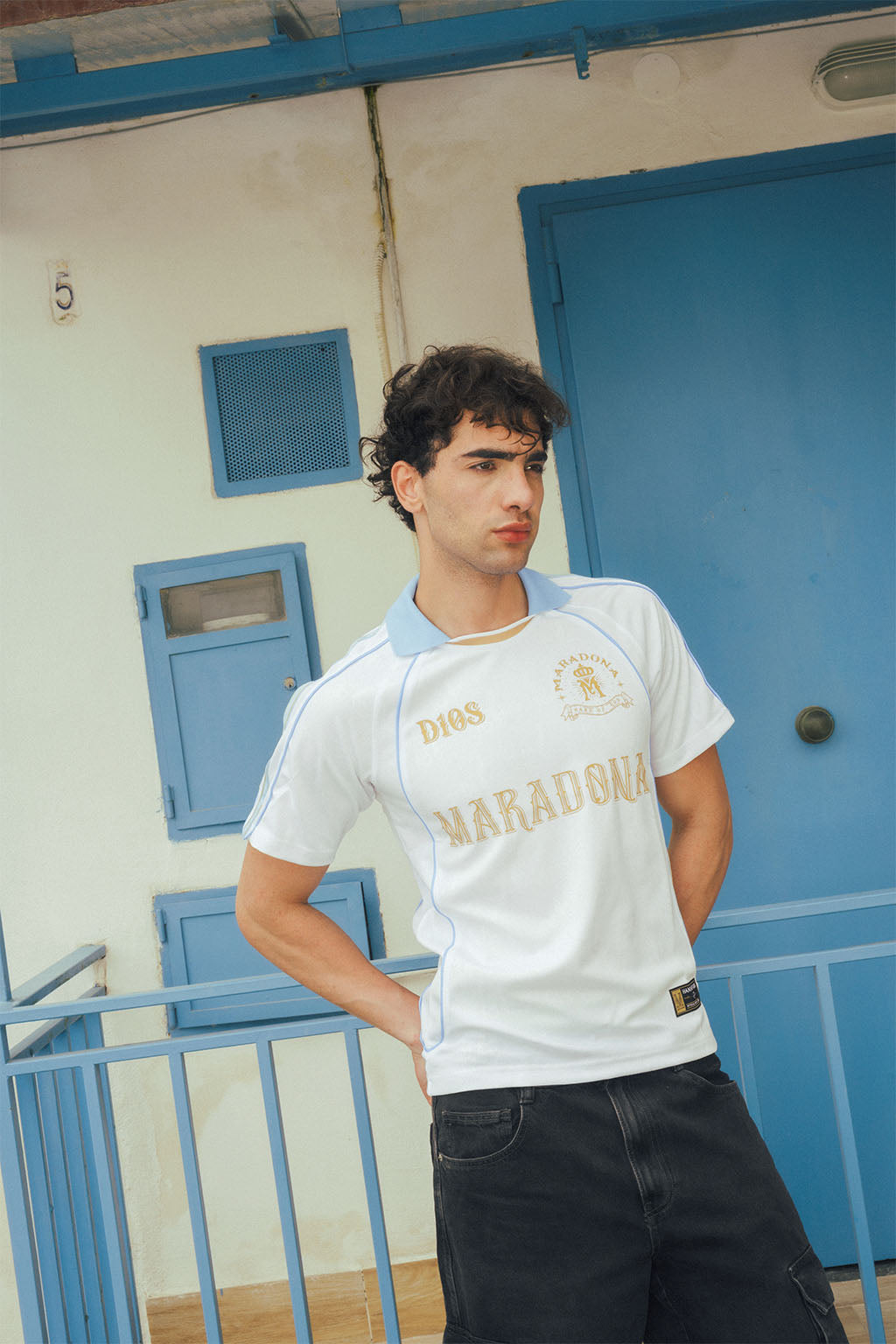 Man wearing a white sports jersey with vintage gold Maradona branding in front of a blue accent building.