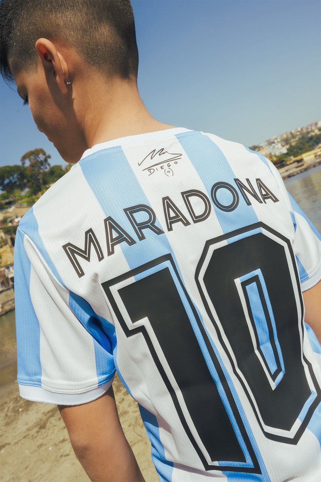 Young boy wearing a sports jersey with 'Maradona 10' on the back, standing on a beach with city background.