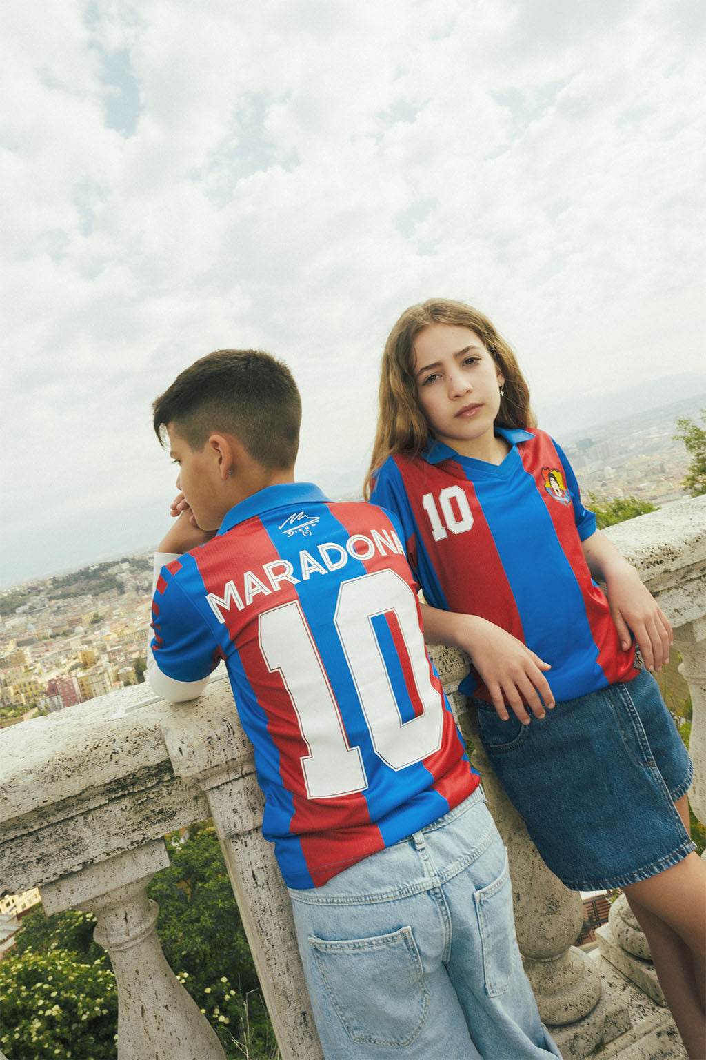 Two children wearing red and blue replica Barcelona jerseys with 'Maradona' and number '10' on a scenic city background.