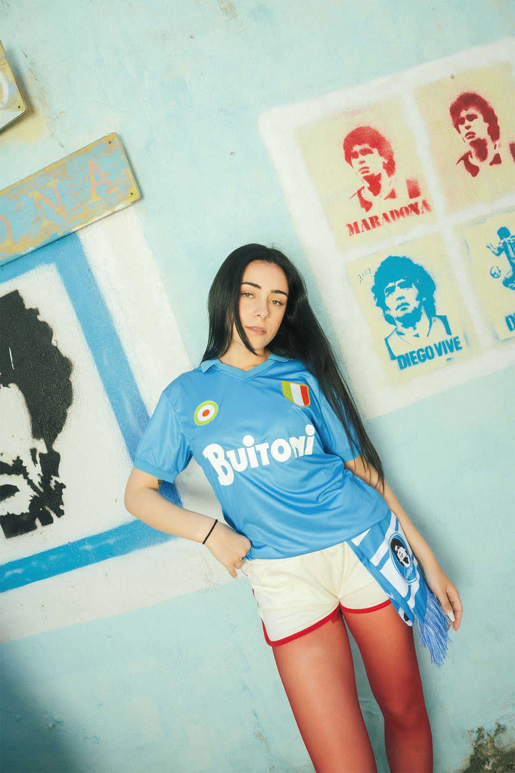 Woman wearing a replica Napoli blue sports jersey posing in front of Maradona posters and blue wall
