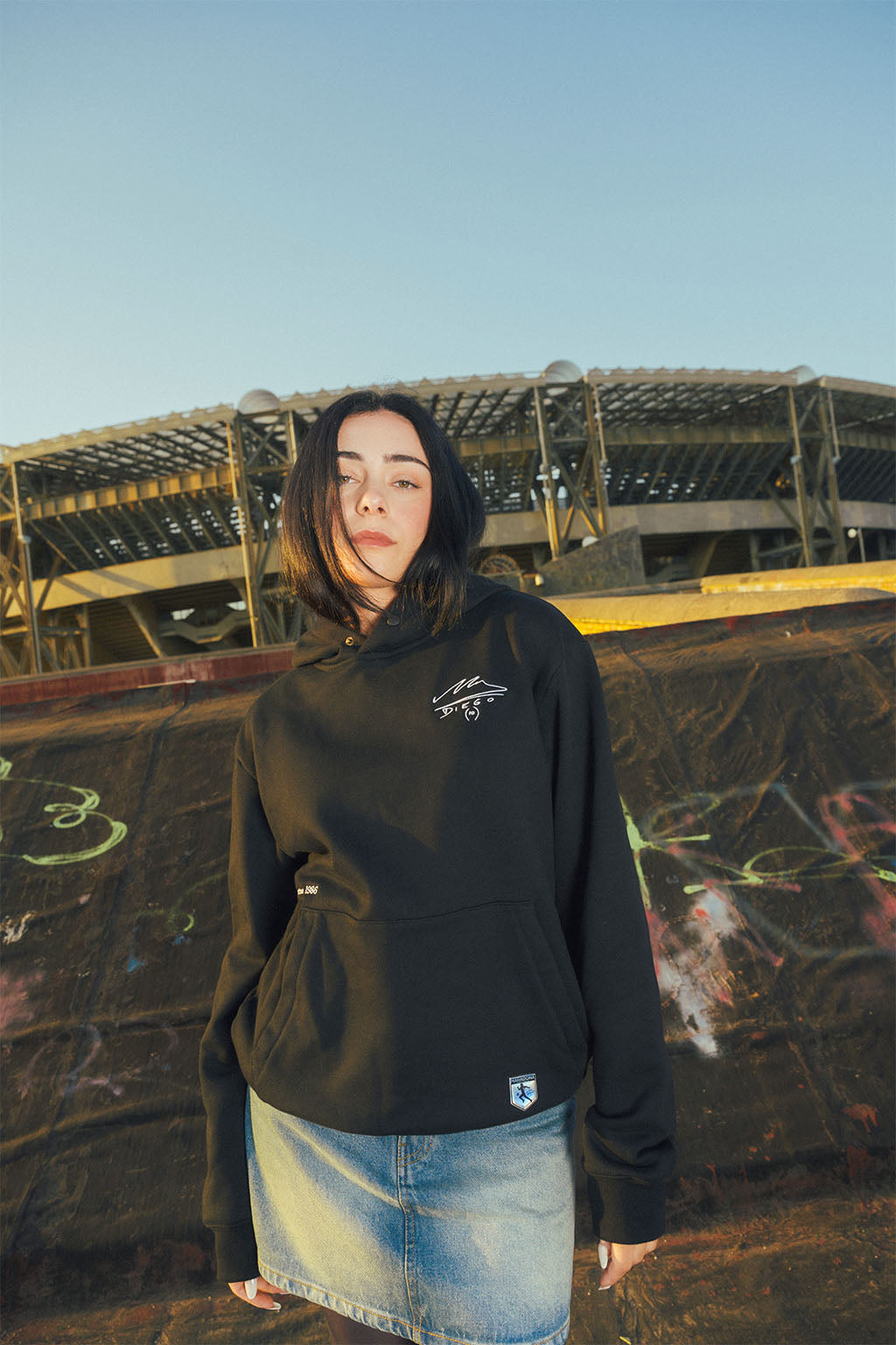 Woman wearing a black hoodie with Maradona signature logo in front of an abandoned stadium.