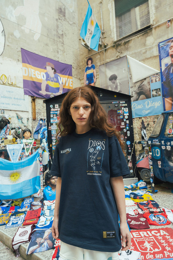 Woman wearing a navy tee with Hand of God 86 graphic print in an urban setting with Maradona flags and posters.