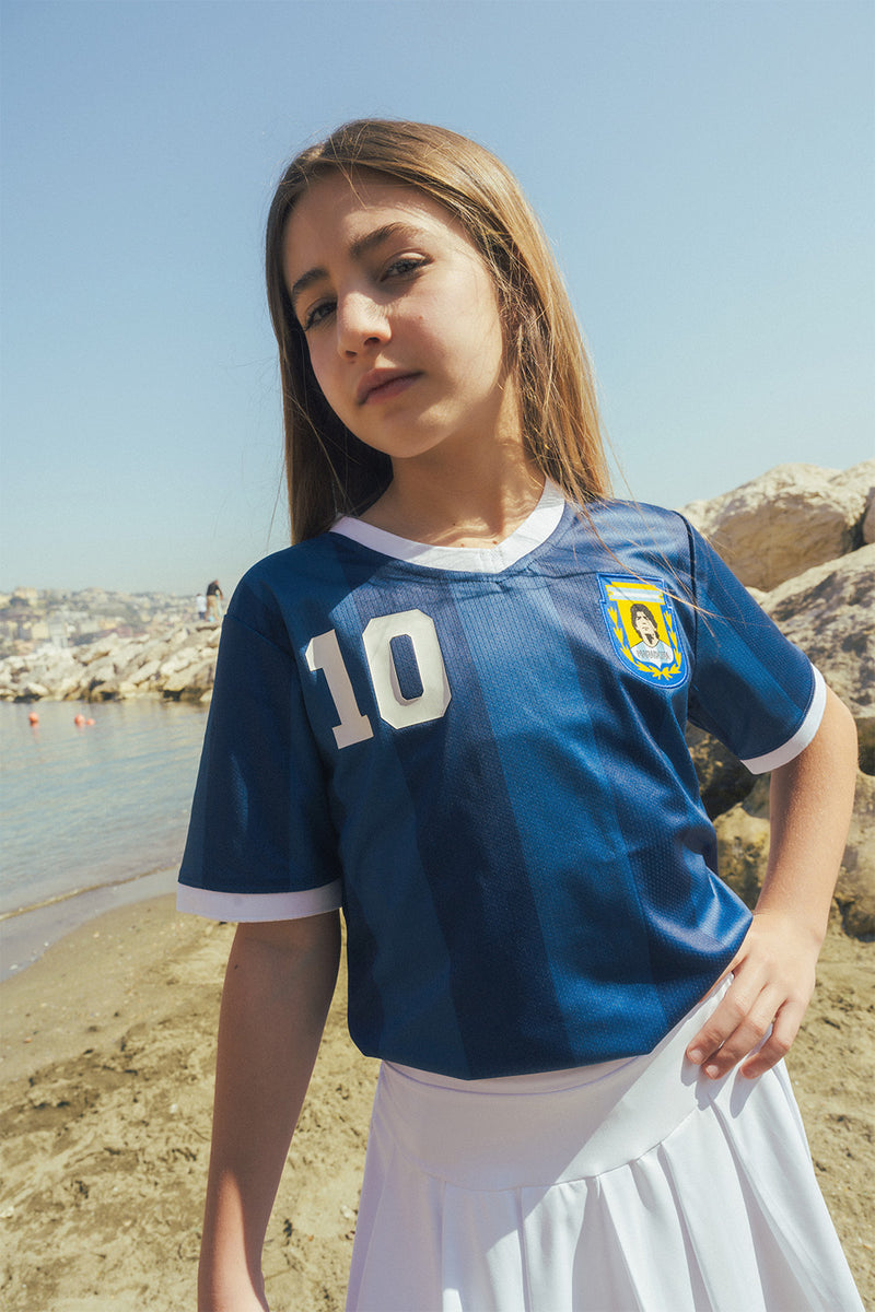 Young girl wearing a navy blue sports jersey with number 10 and a Maradona crest, standing on a beach.