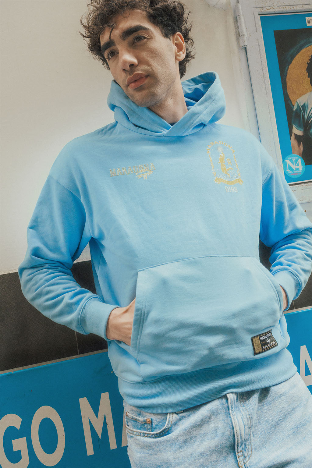 Man wearing a light blue oversize hoodie with gold and white features, standing in front of mural poster.
