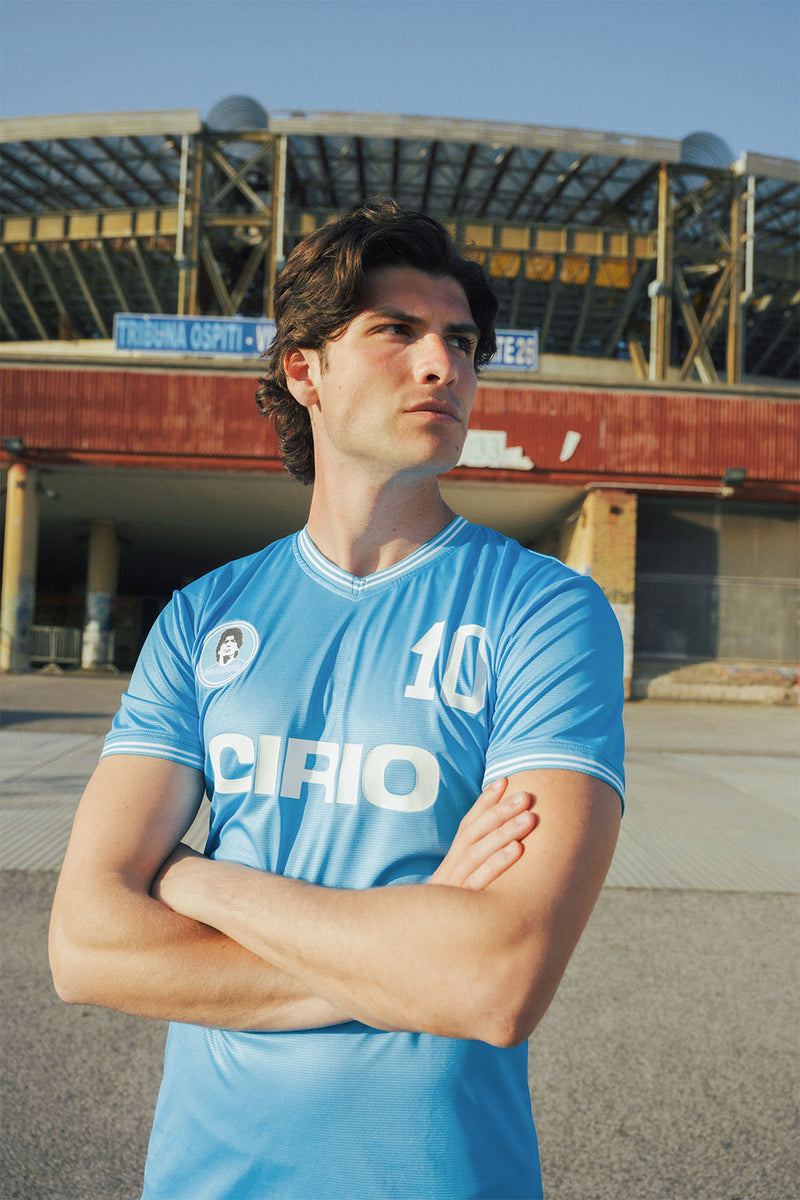 Man wearing a replica Napoli 86 blue sports jersey with Maradona branding outside an old stadium