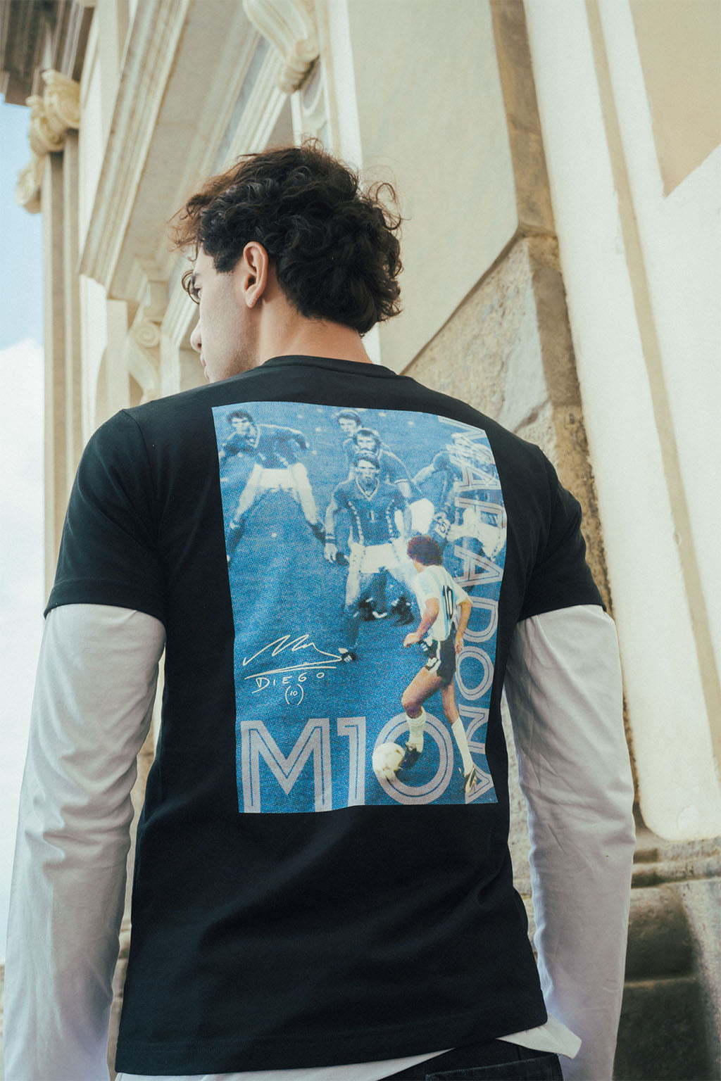 Man wearing a black t-shirt with a blue graphic Maradona 86 design on the back, standing in front of classical architecture.