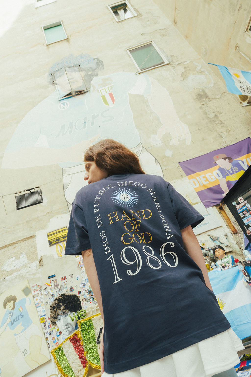 Woman facing away wearing a t-shirt with 'Hand of God 1986' text, standing in front of a Maradona wall art.