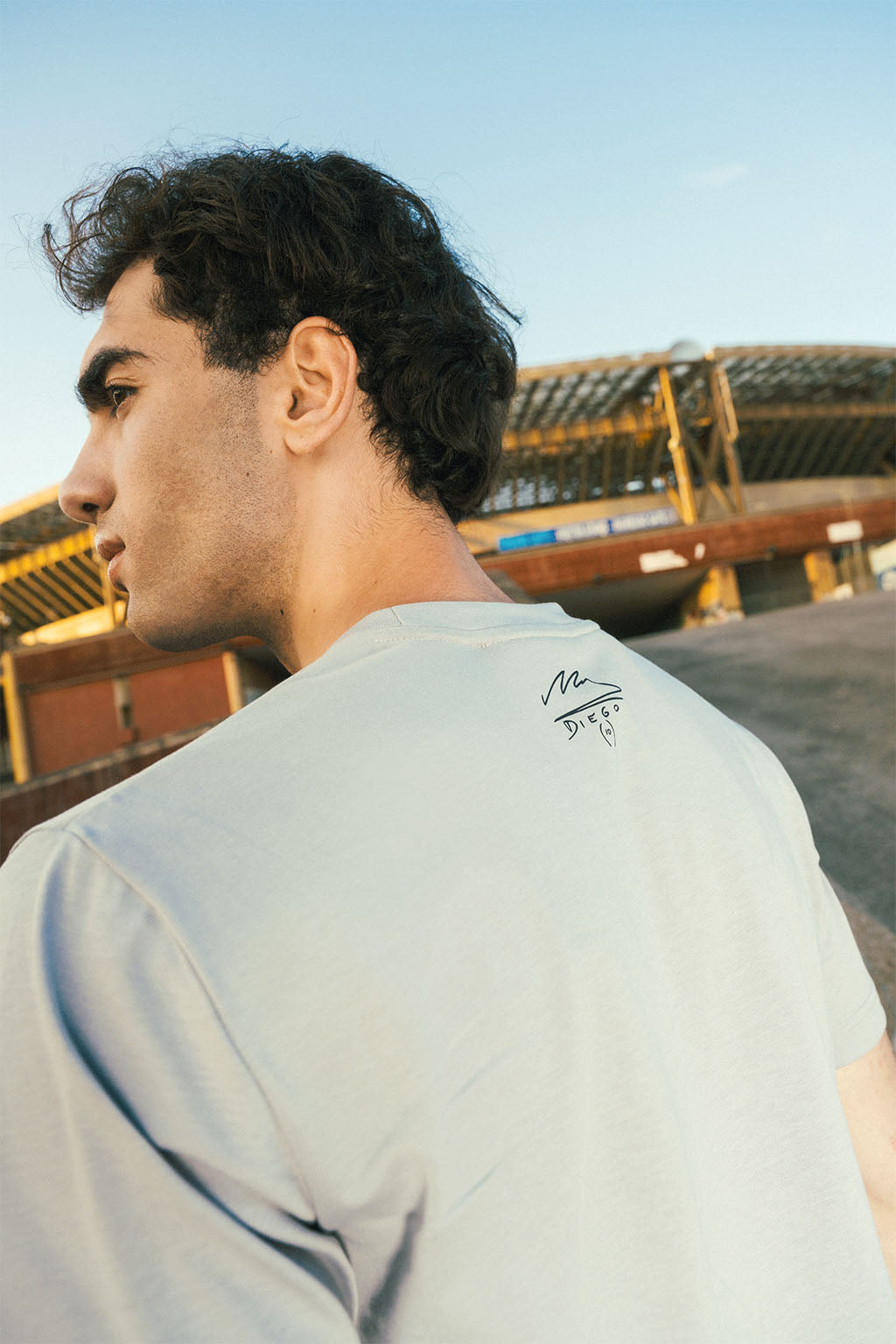 Man wearing an ash gray t-shirt with a Maradona signature logo on the back, standing outside stadium