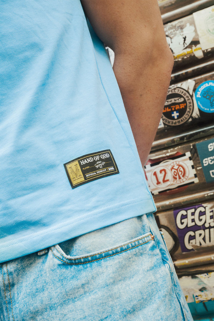 Man wearing a light blue tee with a Hand of God, Maradona brand, standing in front of various stickers.