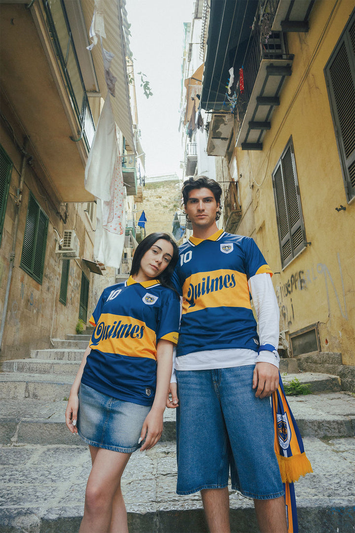 Man and woman leaning against each other wearing matching blue and yellow Boca jerseys in an urban alleyway.
