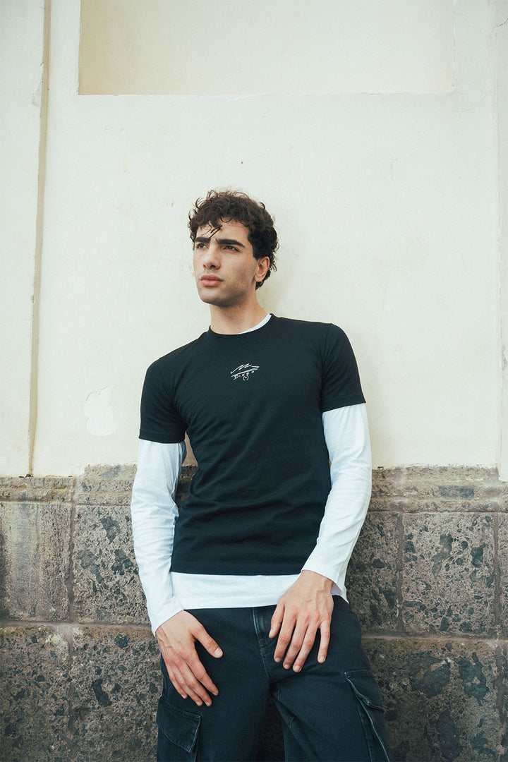 Man wearing a black Maradona signature tee and white long-sleeve under shirt leaning against a wall.
