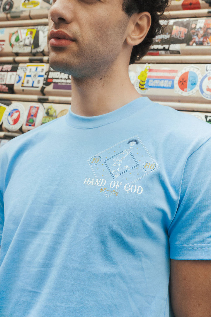 close up of man wearing a light blue t-shirt with 'Hand of God' design in front of a wall of stickers