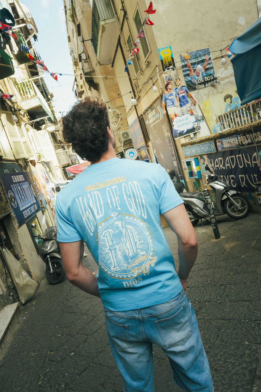 Man wearing a blue t-shirt with a graphic Hand of God design walking down a narrow urban street.