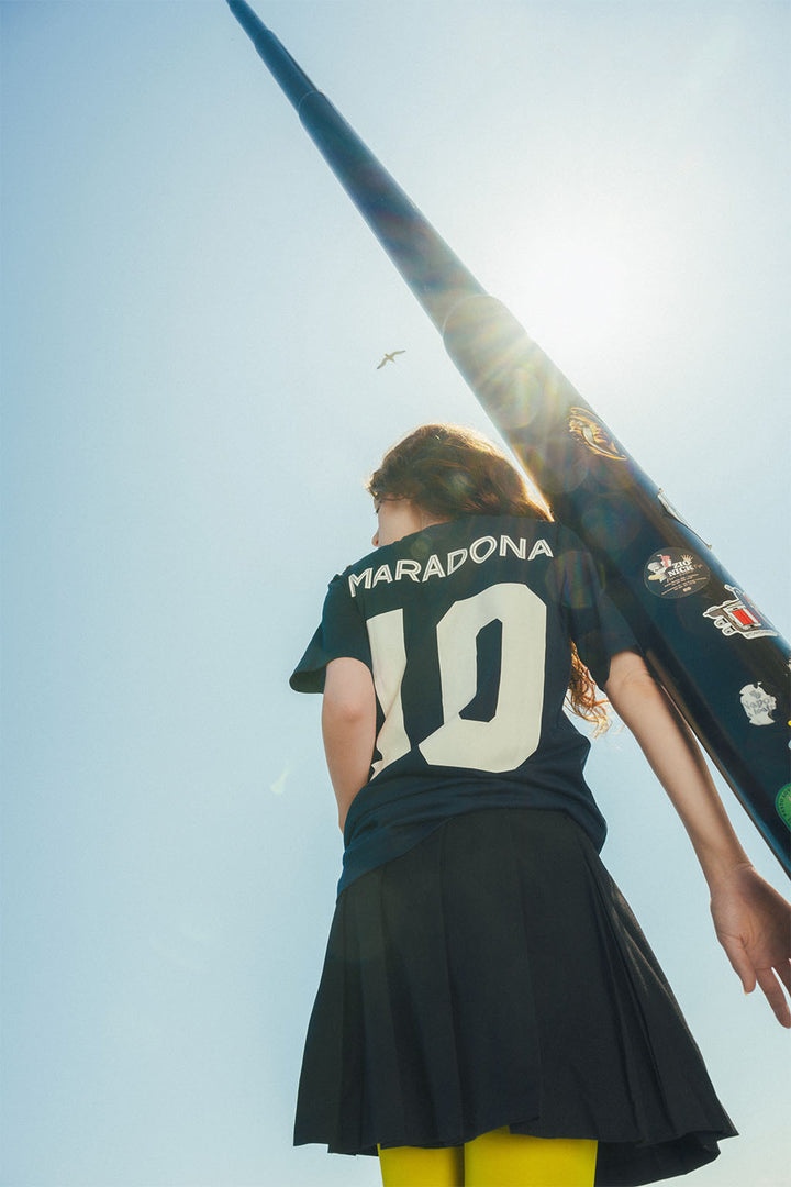 Woman wearing a navy soccer jersey with 'Maradona' and number '10' on back, leaning against a light post under a bright blue sky.