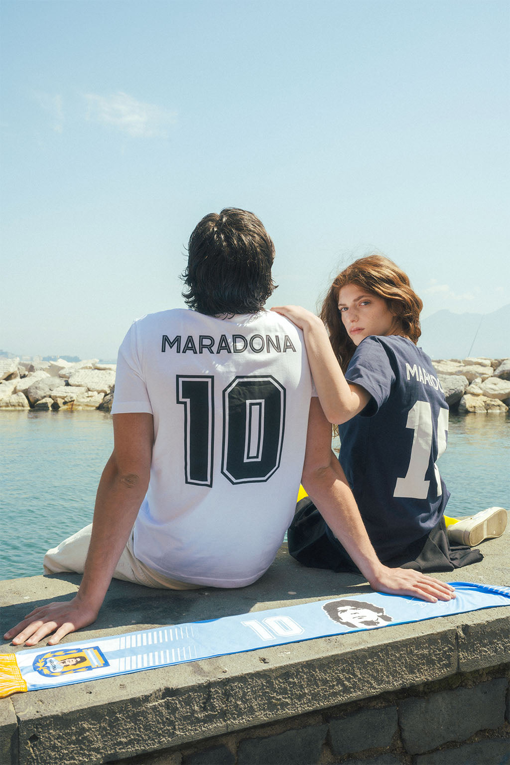 Two people sitting by a waterfront wearing soccer jerseys with 'Maradona' and number '10' on back.