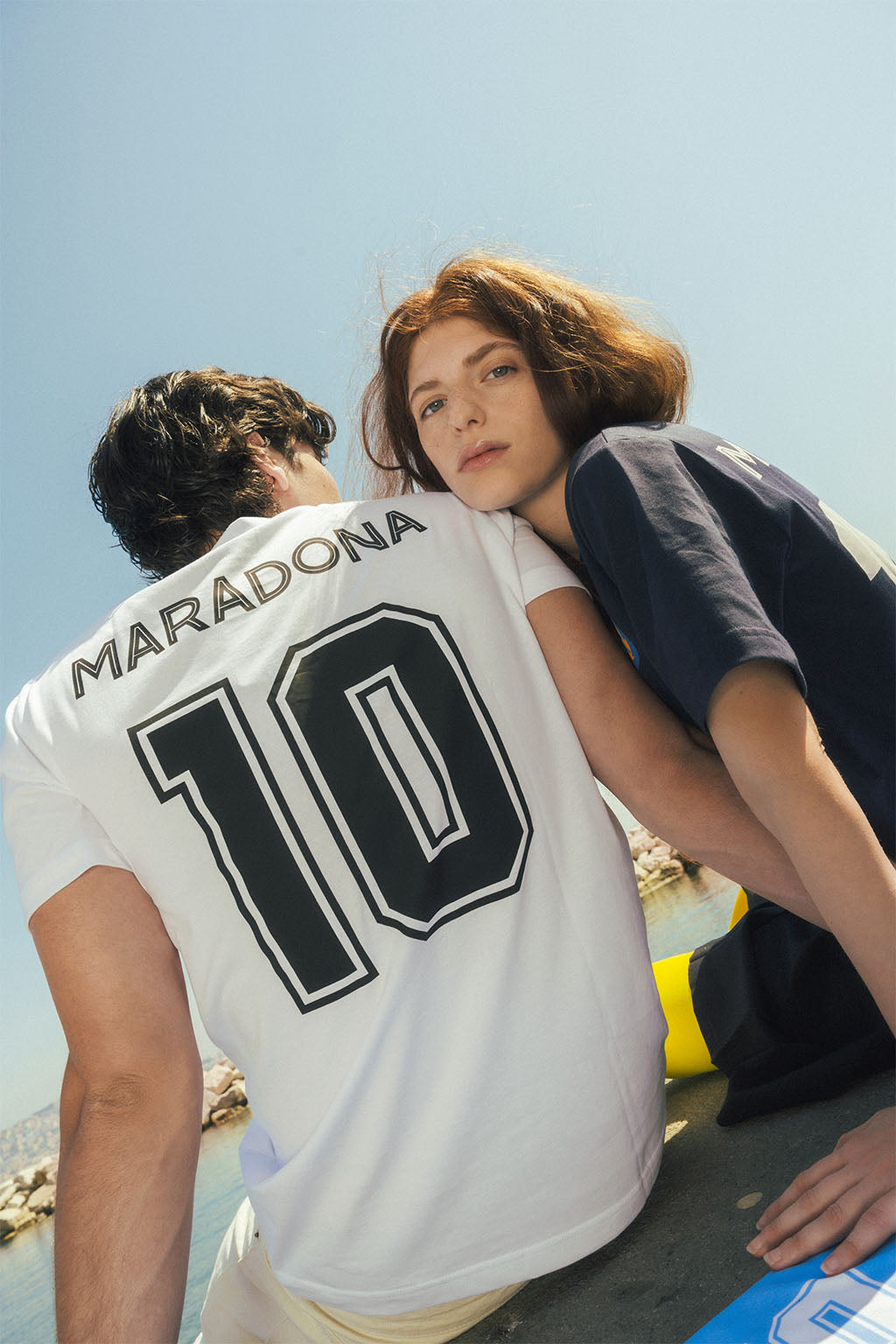 Two people sitting on a beach with one wearing a white jersey with 'Maradona 10' printed on it.