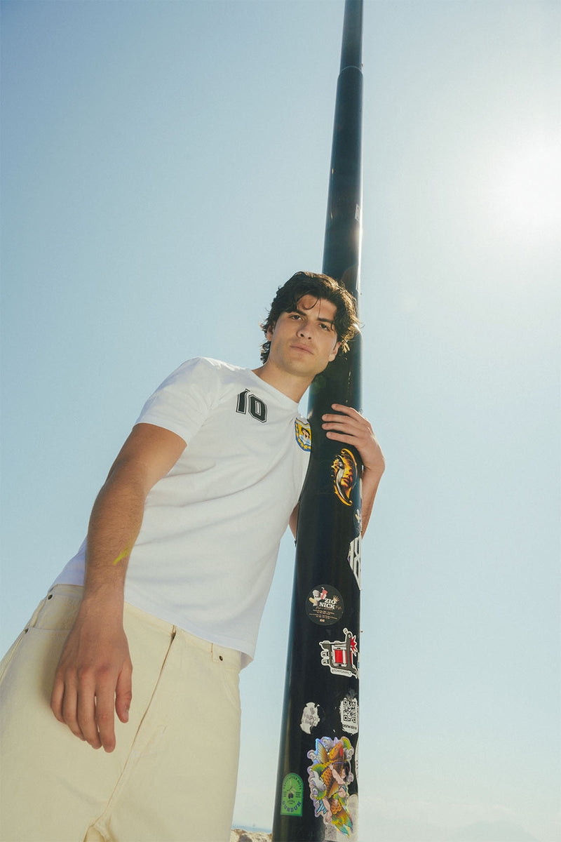 Man leaning against a light post with stickers against a clear blue sky, wearing white Maradona t-shirt